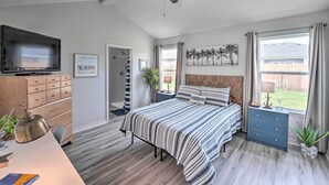 Quarto com cama, cômoda, televisão, ventoinha no teto e janelas com cortinas.