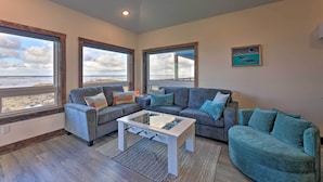 A living room with a gray sofa, a white coffee table, and a view of the beach through large windows. - Sanderling Sea Cottages, Unit 7 w/ Beach Access! (Waldport)