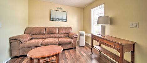 A living room with a brown leather sofa, a wooden coffee table, a wooden side table with a lamp, and a white air conditioner.
