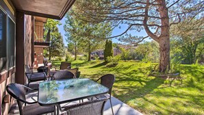 Een terras buiten met een glazen tafel en rieten stoelen, met uitzicht op een verzorgd gazon en bomen.