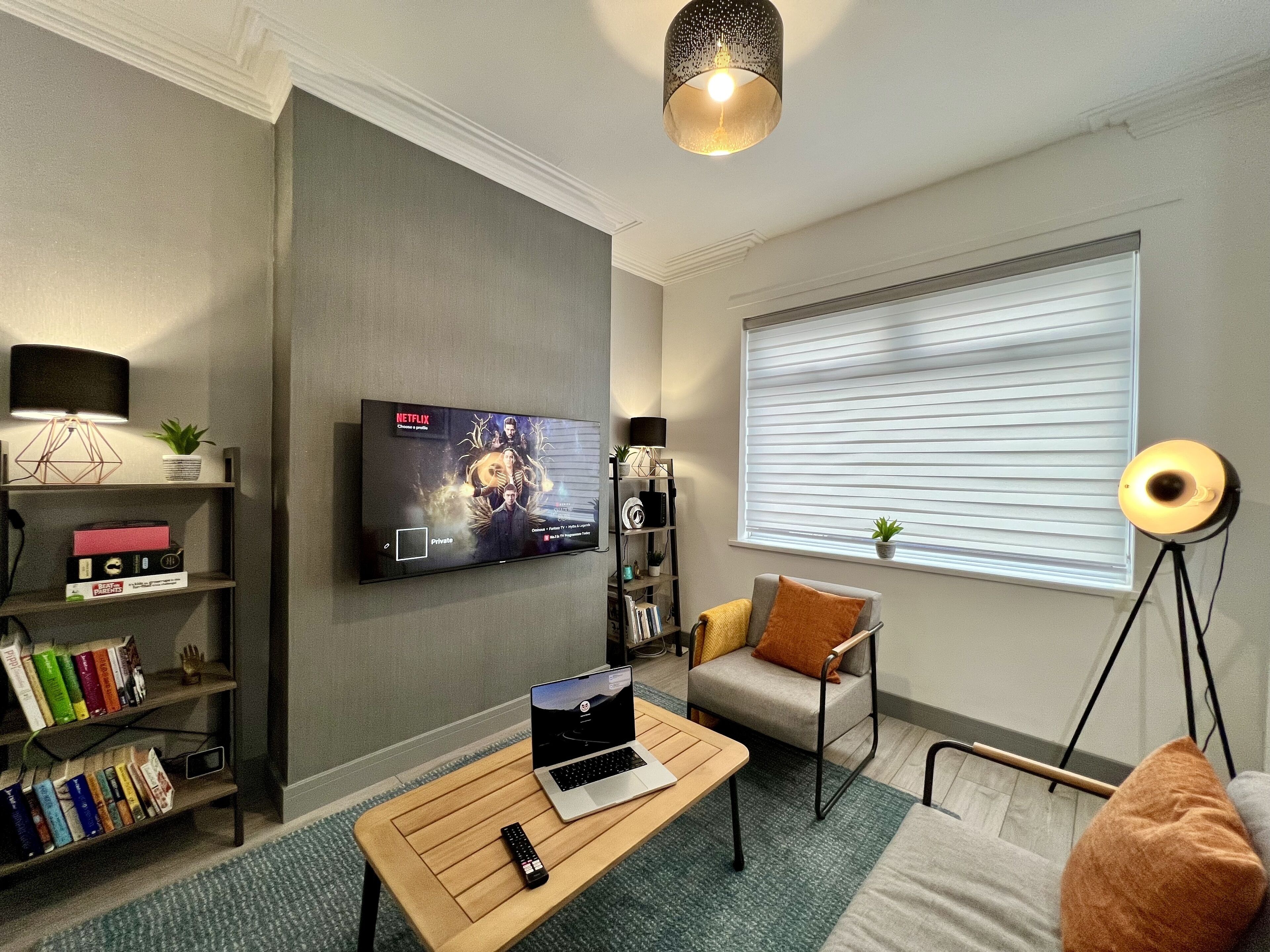 En moderne stue med et fladskærms TV på væggen, en reol, et sofabord med en bærbar computer og en fjernbetjening, en sofa og en gulvlampe.