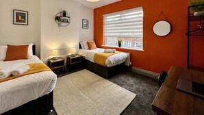 A modern hotel room with two beds, a large window with blinds, a round mirror, and a small table with a laptop. - Daubney House (Cleethorpes)