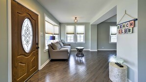 A spacious living room with hardwood flooring, a wooden door with a stained glass window, a beige sofa, a small round table, and a wall-mounted sign reading "BEACH CLUB". - Charming Wildwood Hideaway w/ Private Pool & Deck! (Wildwood)