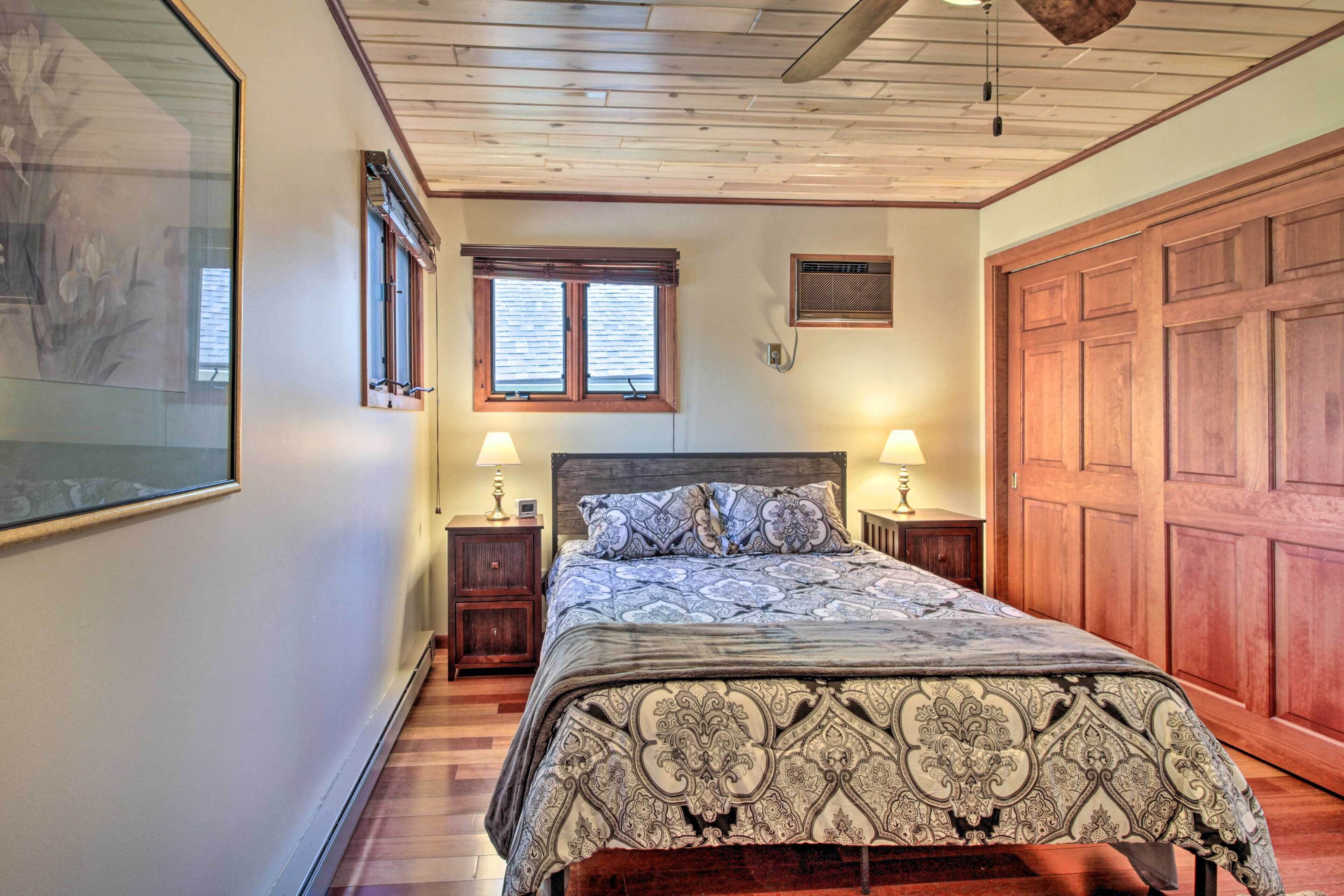 A bedroom with a large bed, wooden ceiling, two bedside tables with lamps, a large wooden door, and a framed picture on the wall.