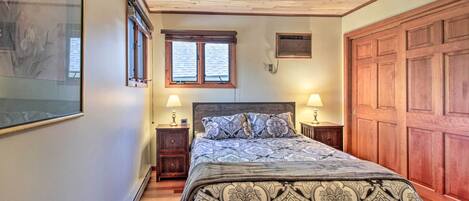 A bedroom with a large bed, wooden ceiling, two bedside tables with lamps, a large wooden door, and a framed picture on the wall.