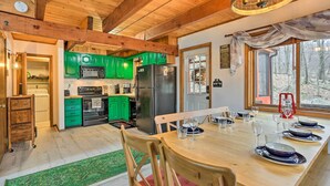 A wooden interior with a dining table set for six, green cabinets, a refrigerator, and a view of a forest through a window. - 4 Mi to Tubing: Cozy Bushkill Cabin w/ Deck! (Bushkill)
