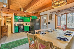 A wooden interior with a dining table set for six, green cabinets, a refrigerator, and a view of a forest through a window.