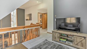 A spacious living area with a large TV, wooden entertainment unit, and a view of the outdoors through a balcony. - 4 Mi to Tubing: Cozy Bushkill Cabin w/ Deck! (Bushkill)