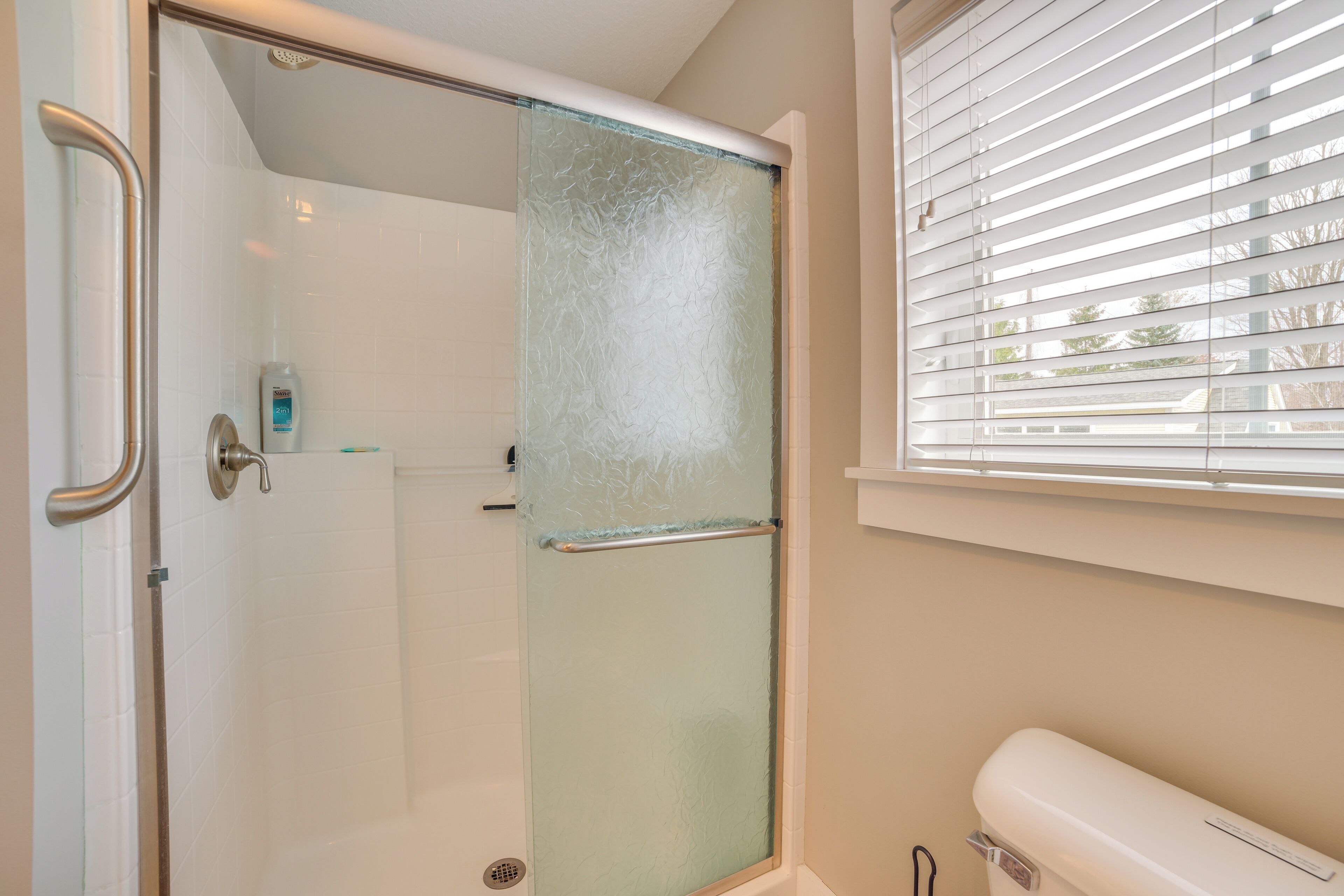 A bathroom with a glass shower door, a toilet, and a window with blinds.