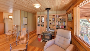 Een gezellig cabin interieur met houten plafond, een houtkachel, een boekenplank en een raam met uitzicht op bomen.