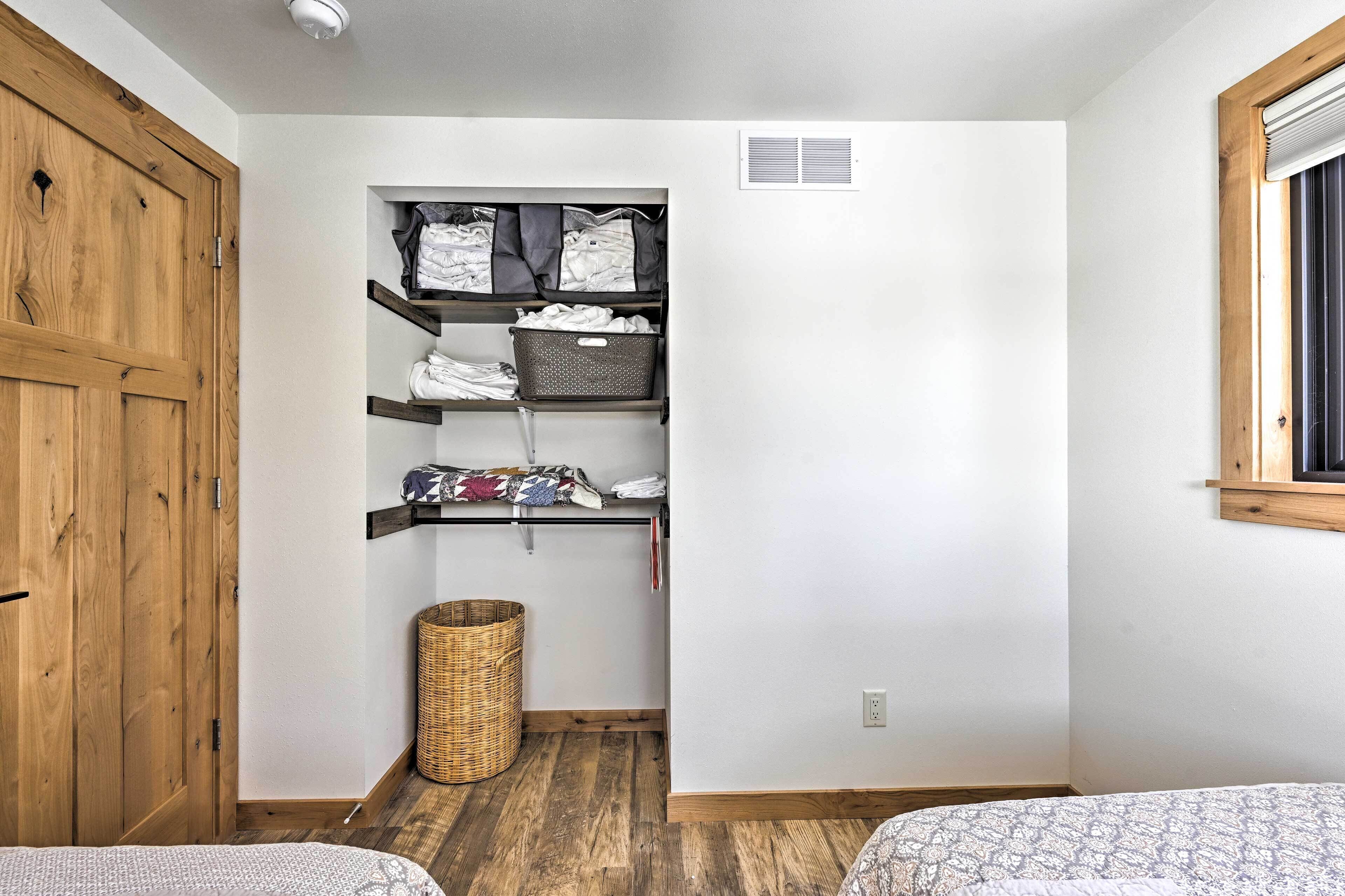 A bedroom with a wooden wardrobe, a built-in shelving unit with folded clothes, a wicker basket, and a window with blinds.