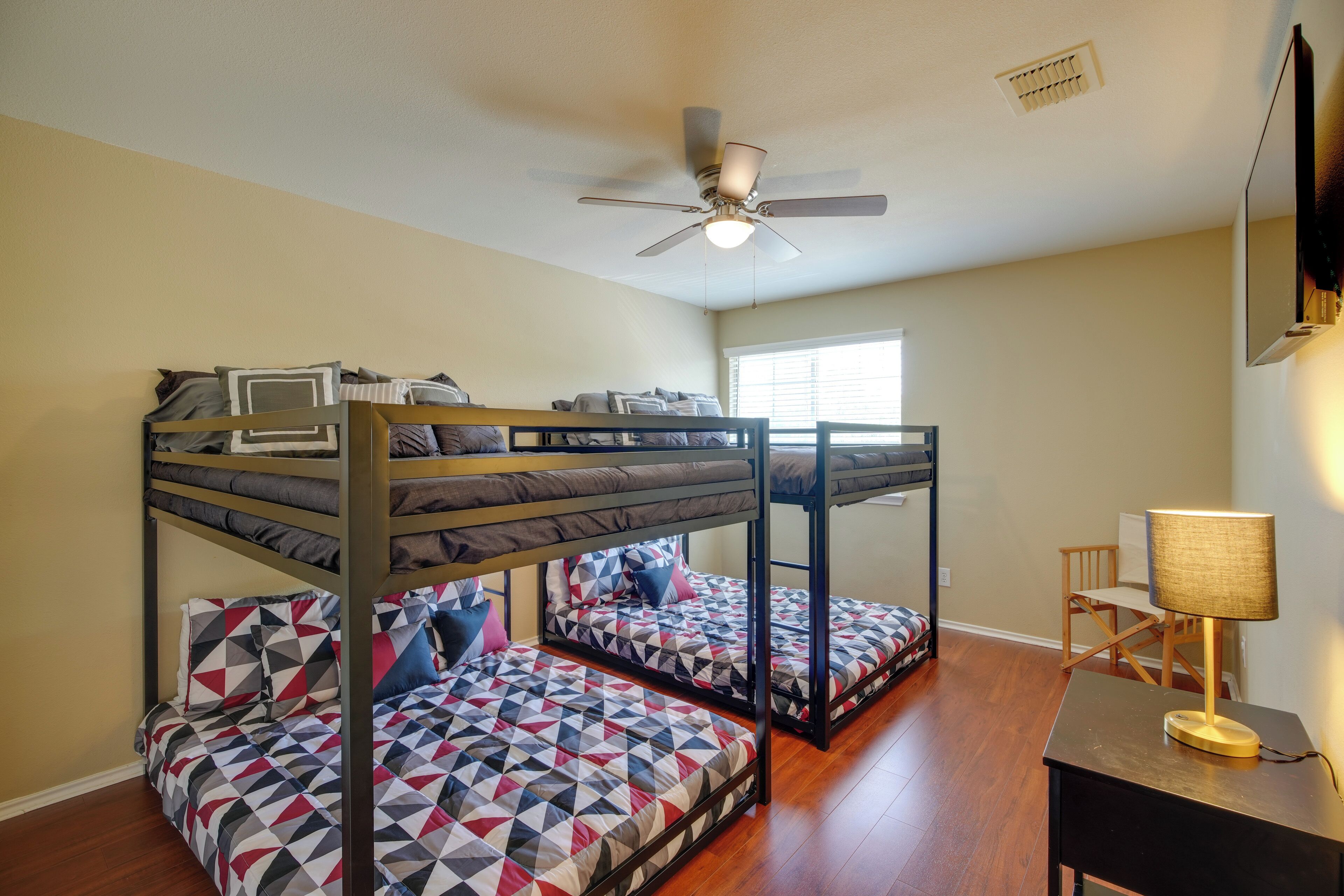 A bunk bed room with a ceiling fan, a desk with a lamp, and a window with blinds.