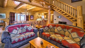 A log cabin interior with a wooden staircase, two patterned sofas, a dining area, and a chandelier. - Exquisite Mccall Log Cabin - Walk to Payette Lake! (McCall)