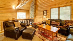 A cozy log cabin interior with a fireplace, leather furniture, and a large window with blinds. - Exquisite Mccall Log Cabin - Walk to Payette Lake! (McCall)