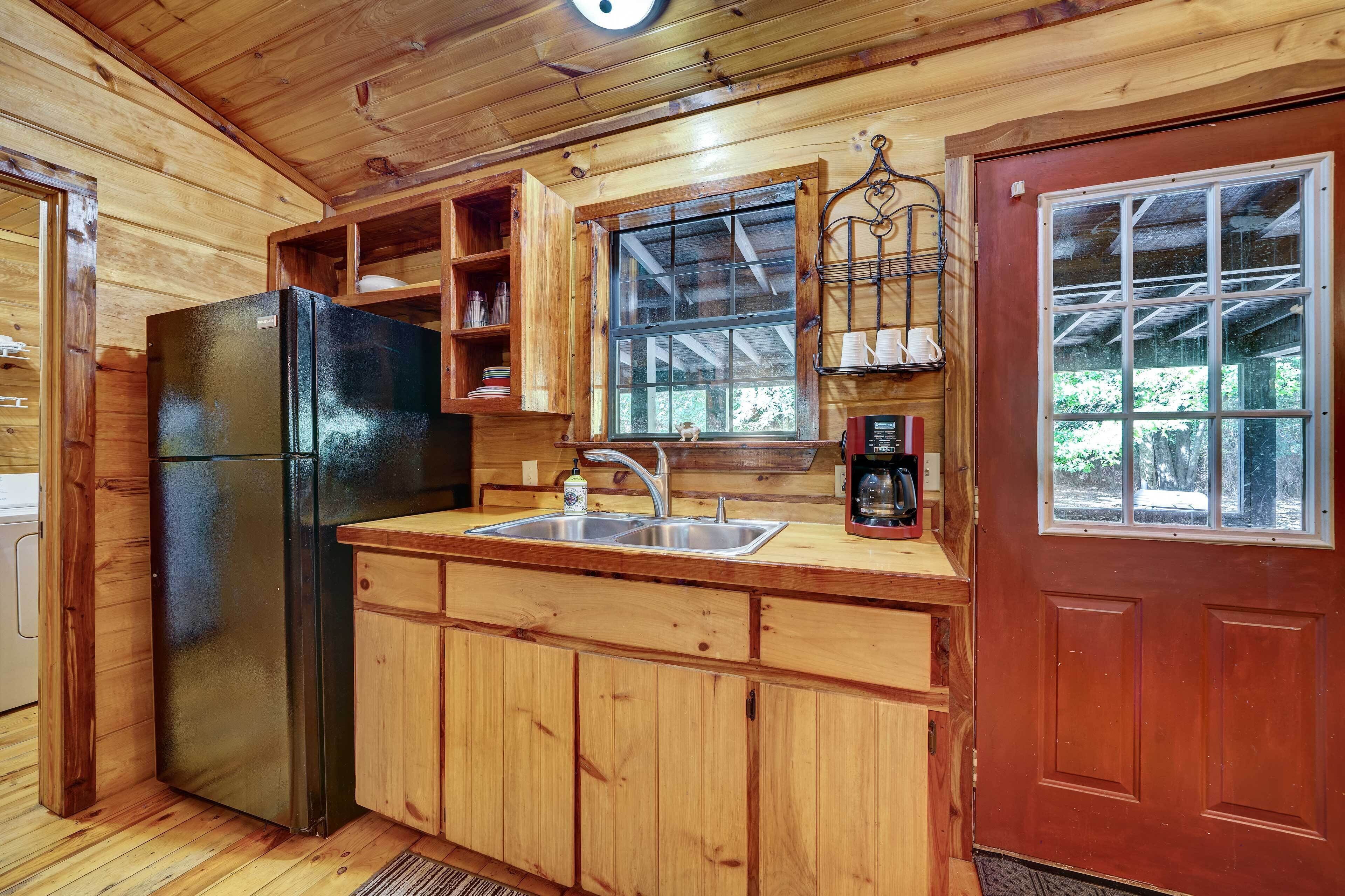 A wooden cabin kitchen with a refrigerator, sink, cabinets, and a window overlooking greenery.