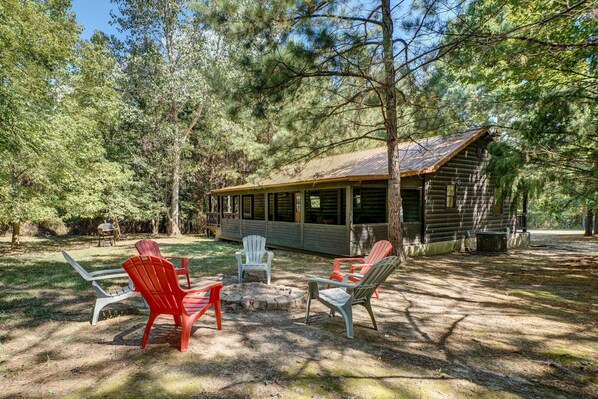 A rustic cabin with a porch, surrounded by trees. There are Adirondack chairs arranged around a fire pit. - Private Broken Bow Cabin w/ Hot Tub + Gazebo! (Broken Bow)