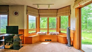 A cozy room with a large window, wooden seating, a flat-screen TV, and a fan. - Scenic Bartlett Home w/ Deck - 1 Mi to Storyland! (Glen)