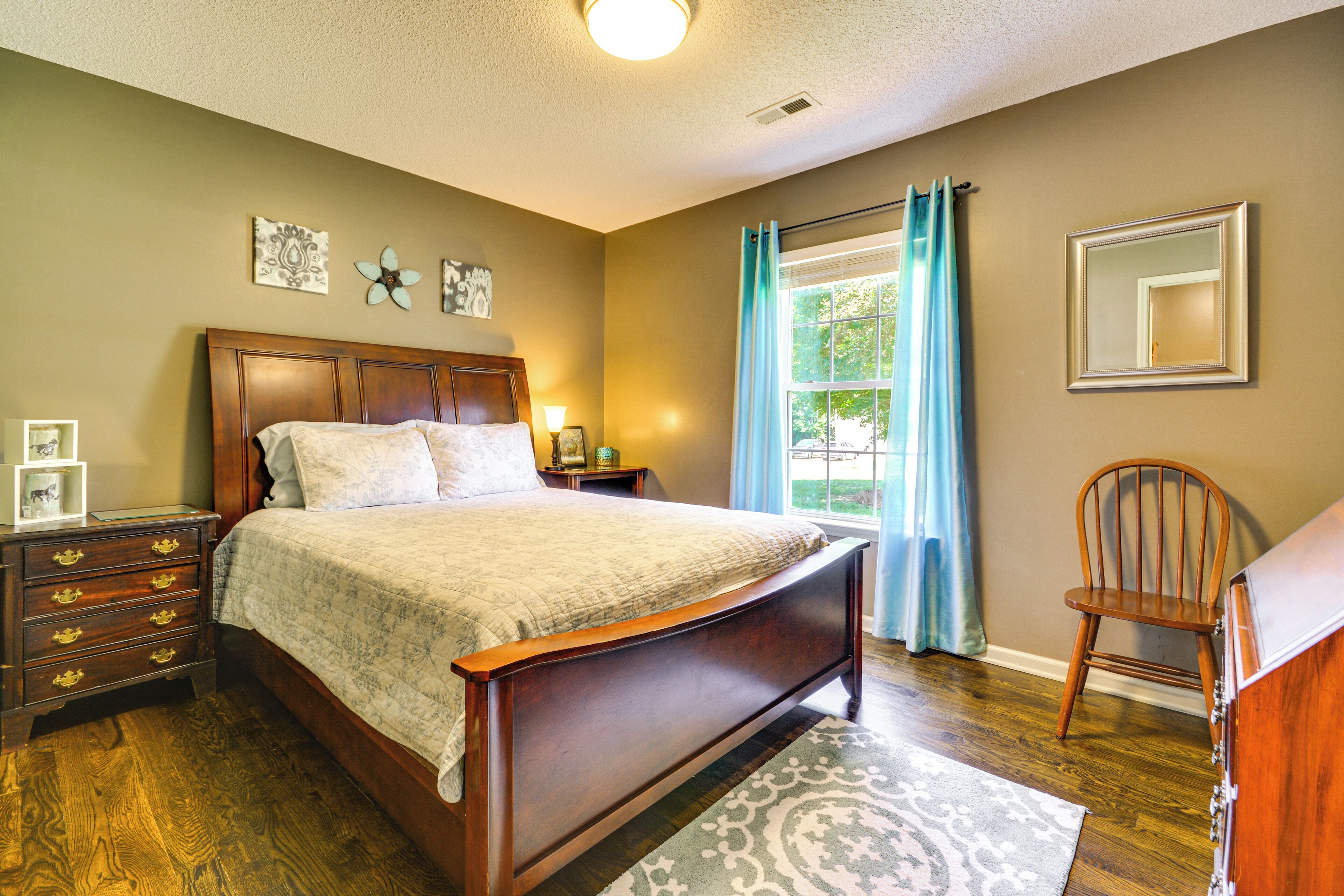 Une chambre à coucher avec un lit en bois, une table de chevet, une commode, une chaise et une fenêtre avec des rideaux.