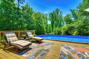 A wooden deck with two lounge chairs and a swimming pool surrounded by trees.