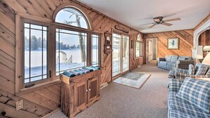 Een gezellig interieur van een chalet, met grote ramen, een houten plafond, een plafondventilator en een geruit bankje.