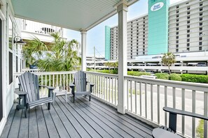 A covered outdoor area with two Adirondack chairs and a small table, overlooking a parking area and a tall building with a sign. - Luxury Home w/ Ocean View, Private Pool, & Hot Tub (Murrells Inlet)