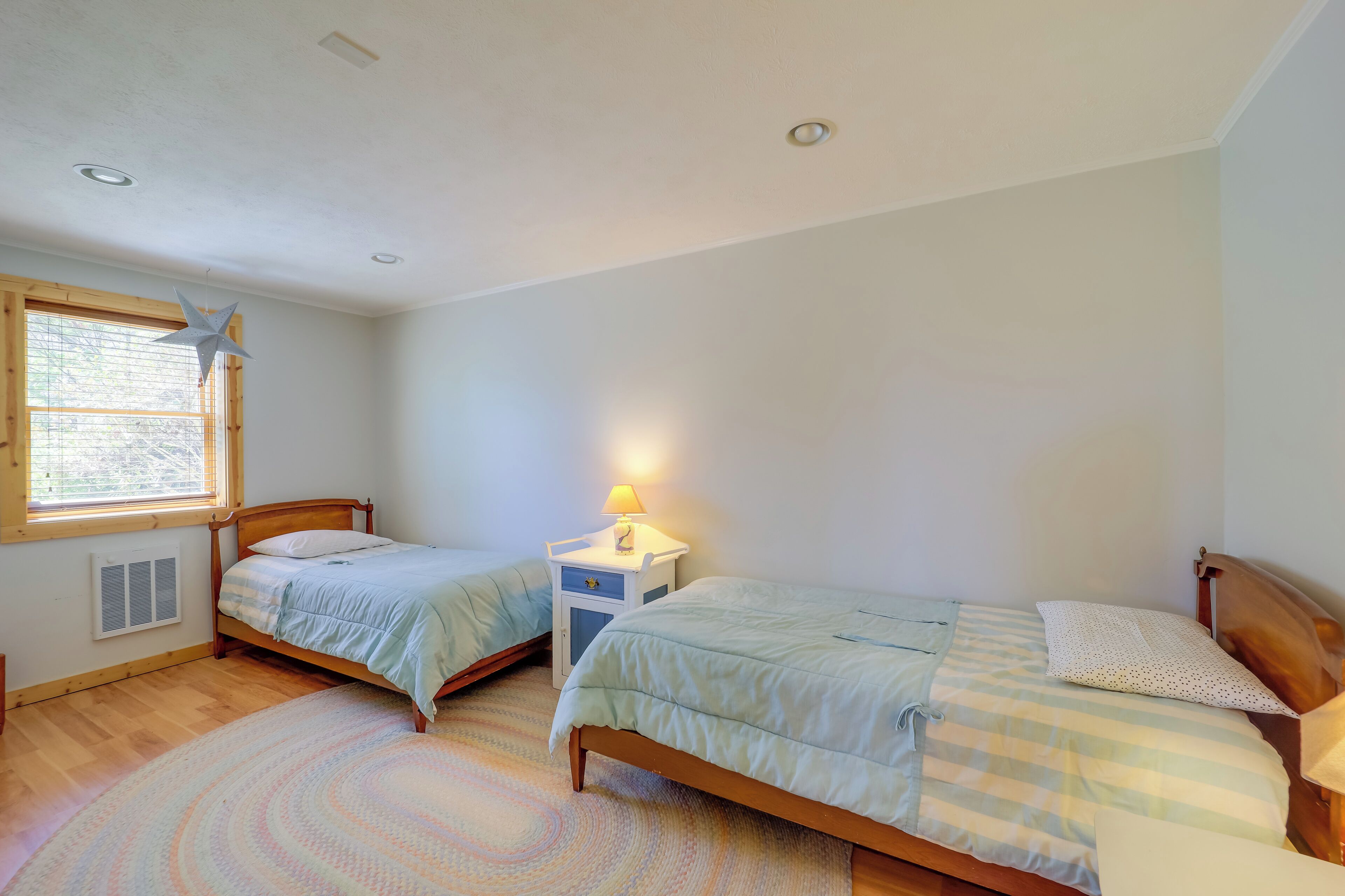 A bedroom with two single beds, a window with blinds, a bedside table with a lamp, and a patterned rug.