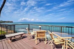 A wooden deck with a bench and two chairs overlooking a beach with clear blue water and a distant shoreline.