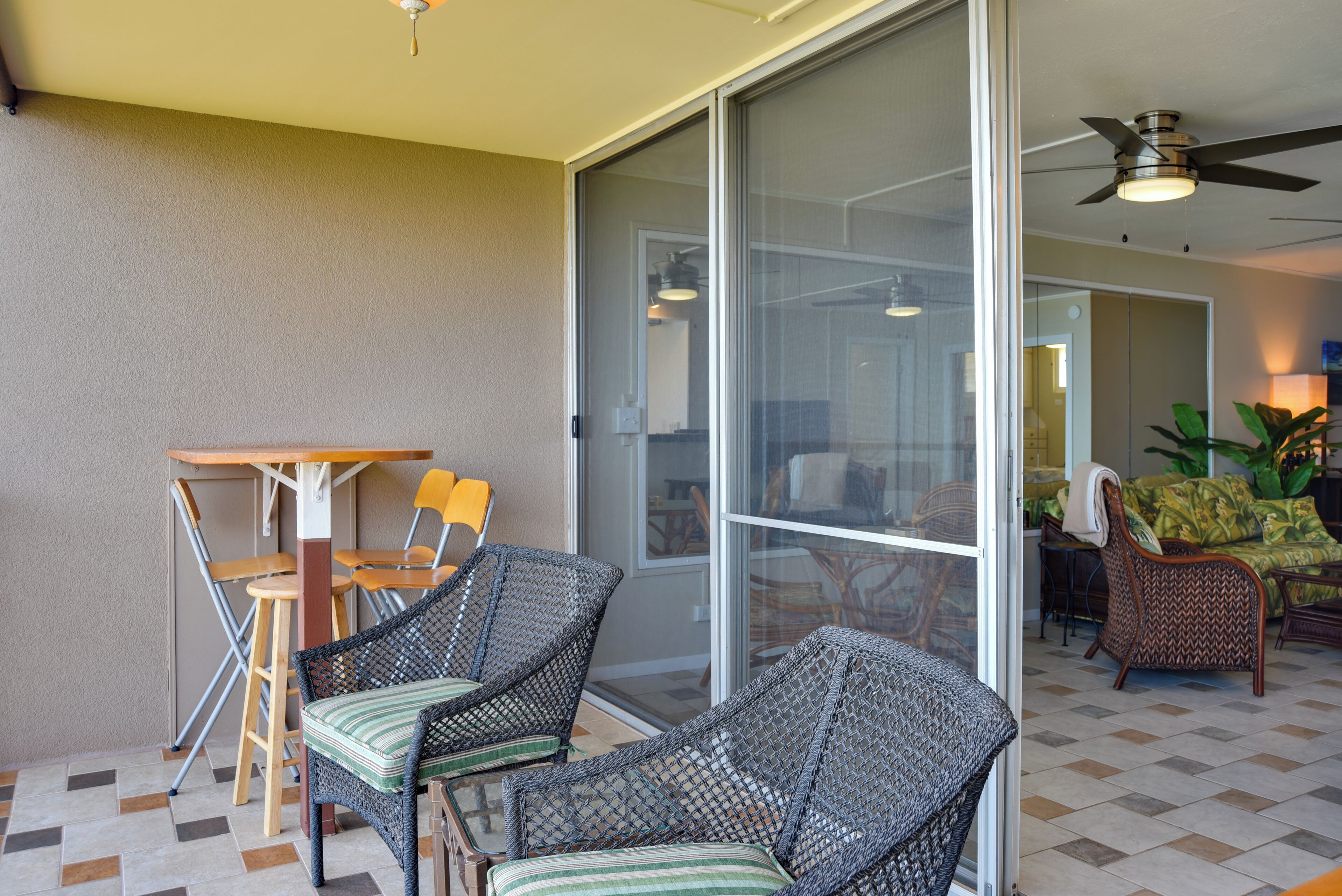 A room with a sliding glass door leading to a balcony, wicker chairs, a small table, and a ceiling fan.