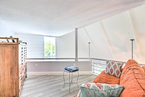 A spacious room with a high ceiling, a wooden cabinet, a small round table with a book, a sofa with cushions, and a window with a view of greenery.