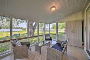 A covered patio with wicker furniture, a glass table, and a view of a lake and grassy area.