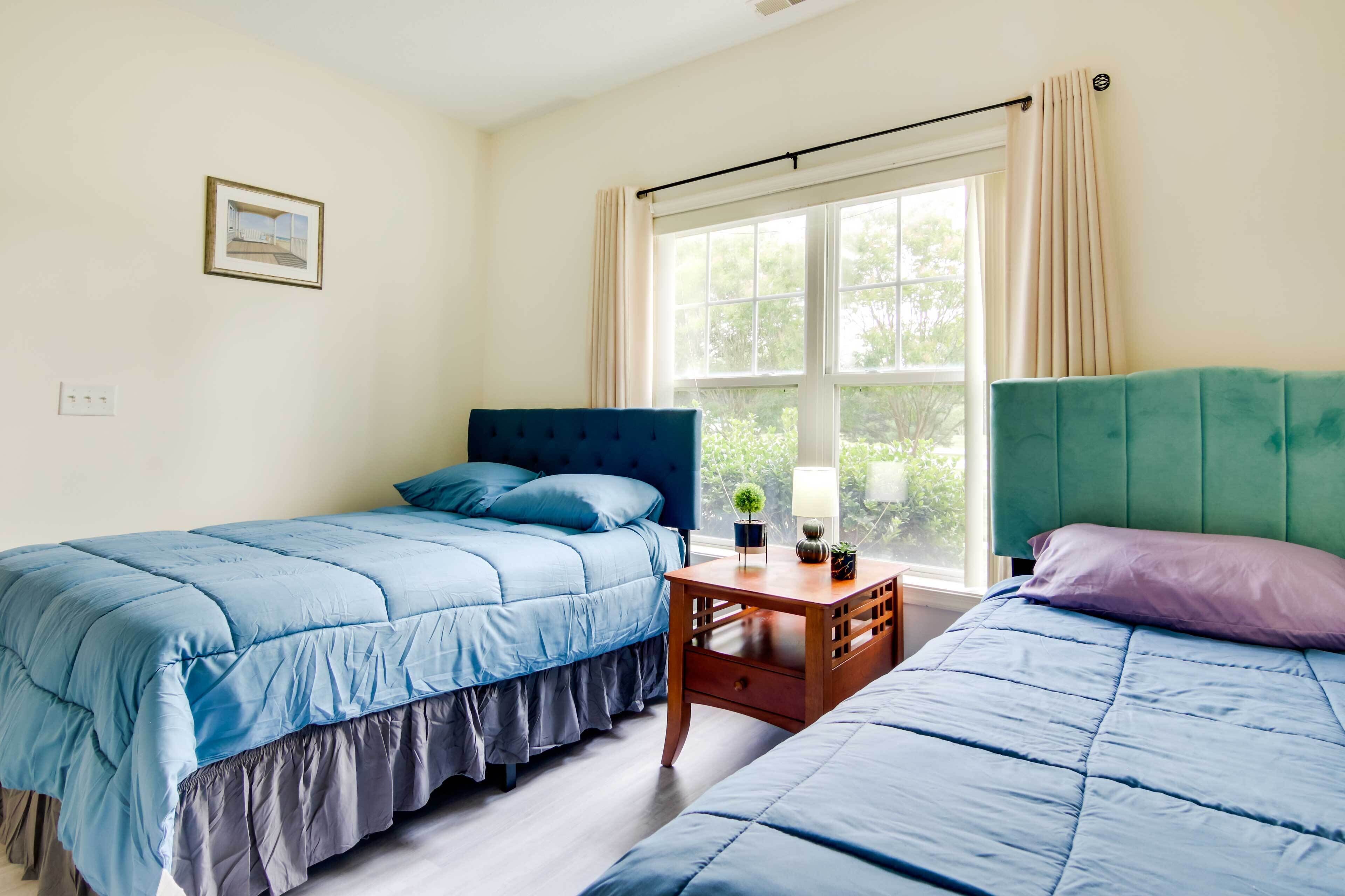A bedroom with two beds, a window with curtains, a bedside table with a lamp, and a framed picture on the wall.