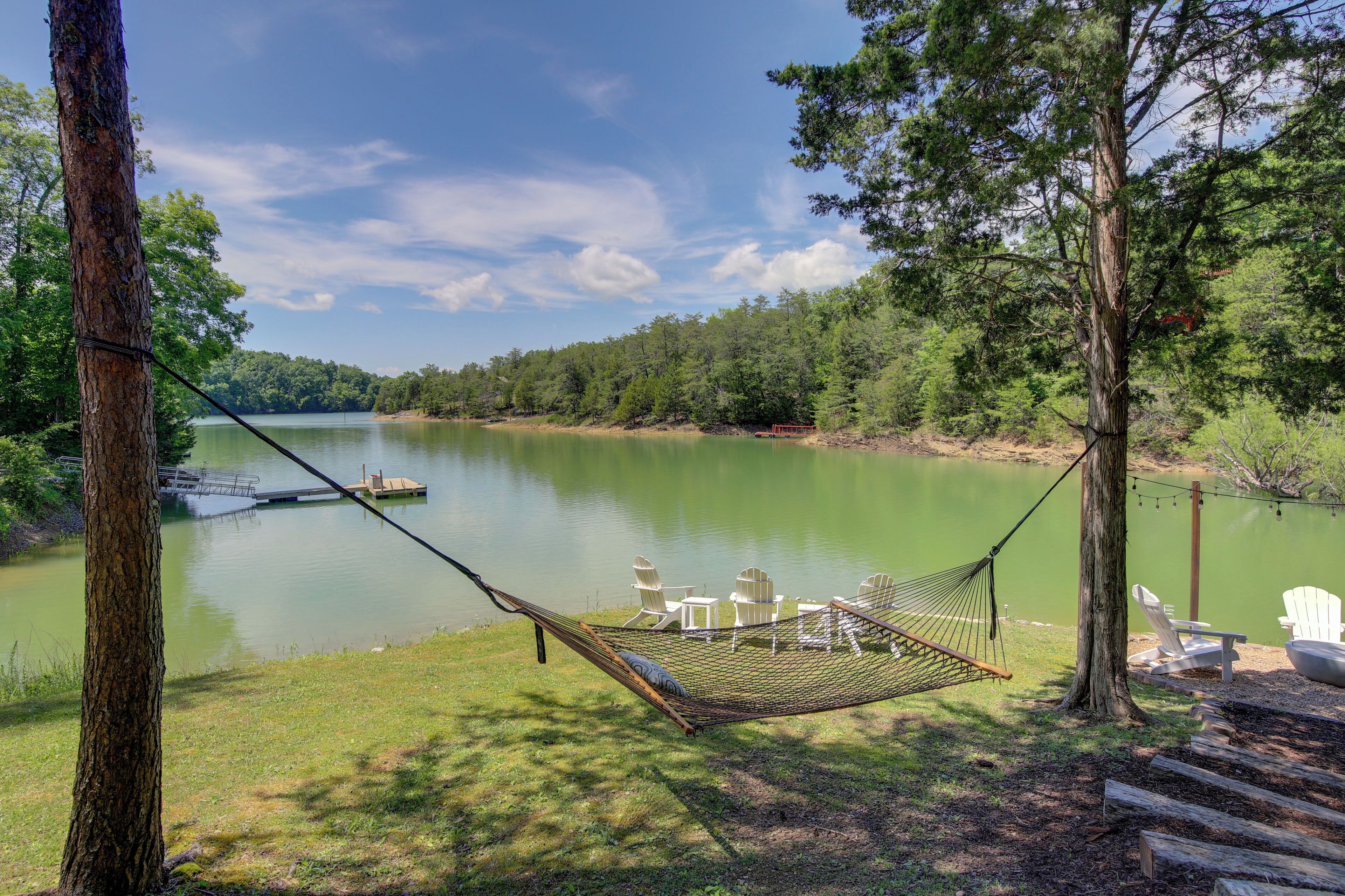 A hammock with chairs by a calm lake, surrounded by trees.