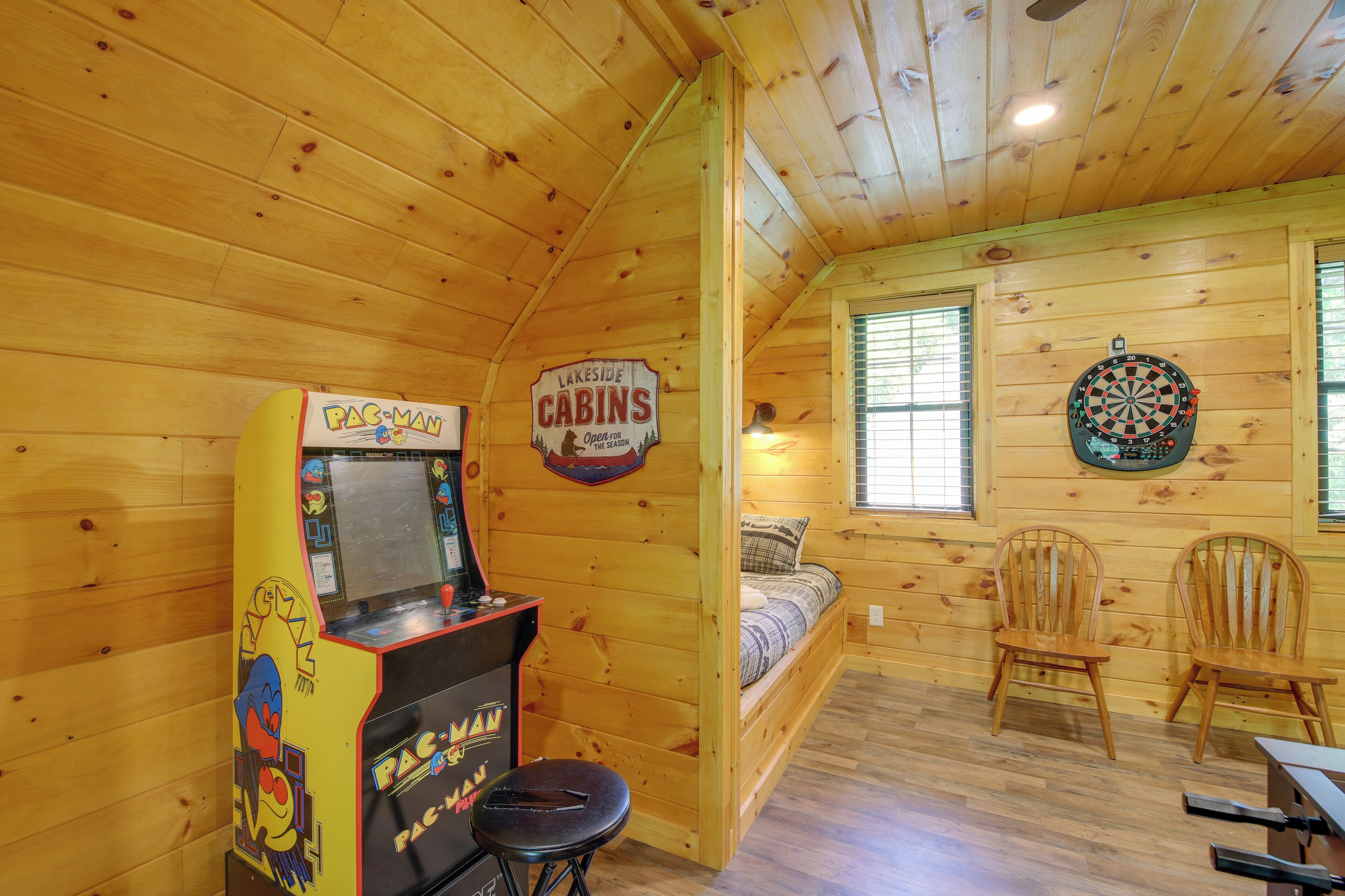 A room with wooden walls and a bed, a Pac-Man arcade machine, and a dartboard.