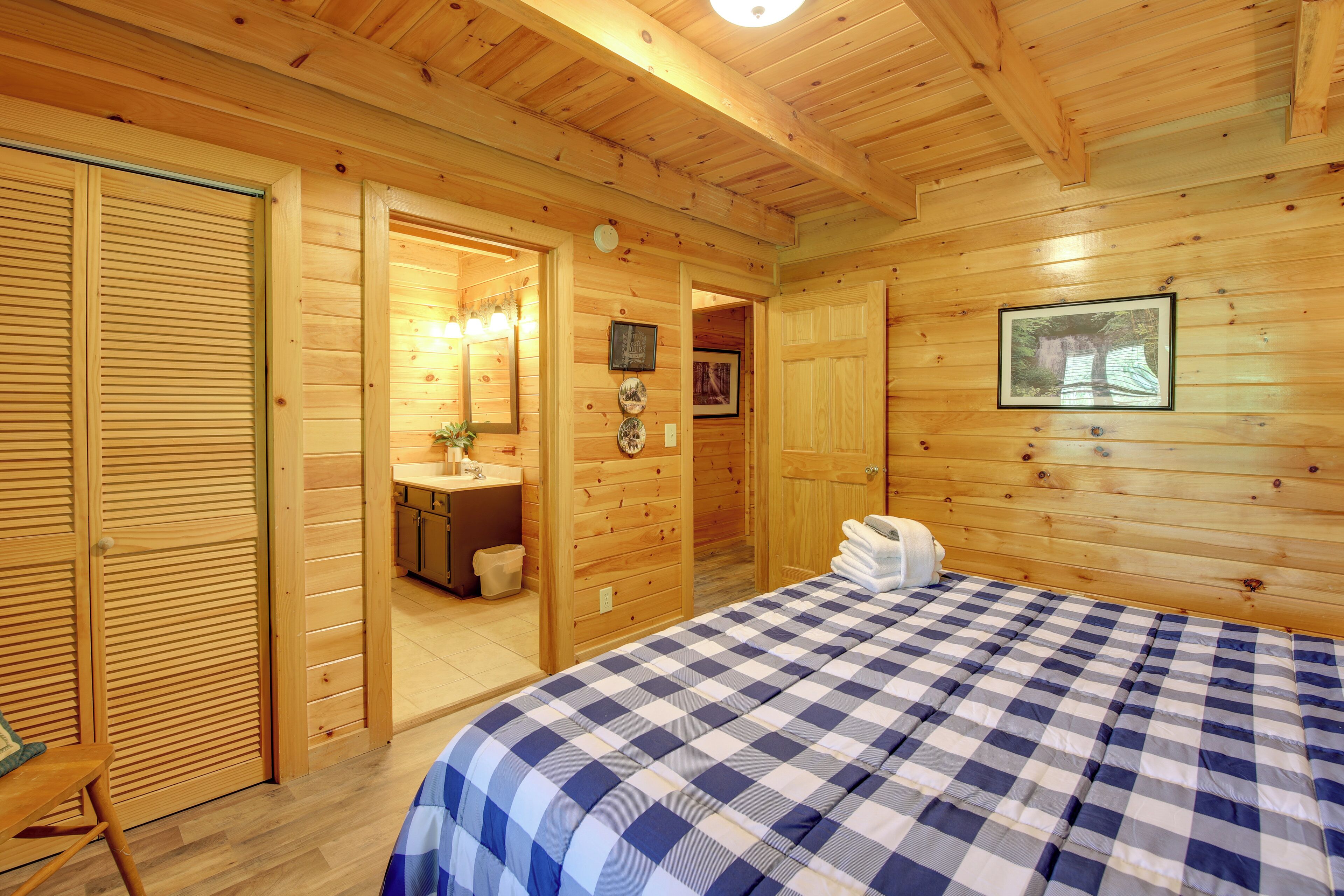 Une chambre de style cabane en bois, avec une couvre-lit à carreaux, un plafond en bois, une salle de bain dotée d’un lavabo et d’un miroir, et un tableau encadré accroché au mur.