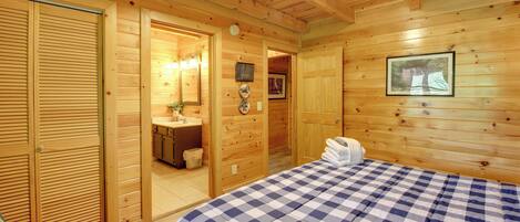 Ein Zimmer in einer Holzhütte mit karierter Bettdecke, Holzboden und einem Badezimmer mit Waschbecken und Spiegel. Ein gerahmtes Bild hängt an der Wand.