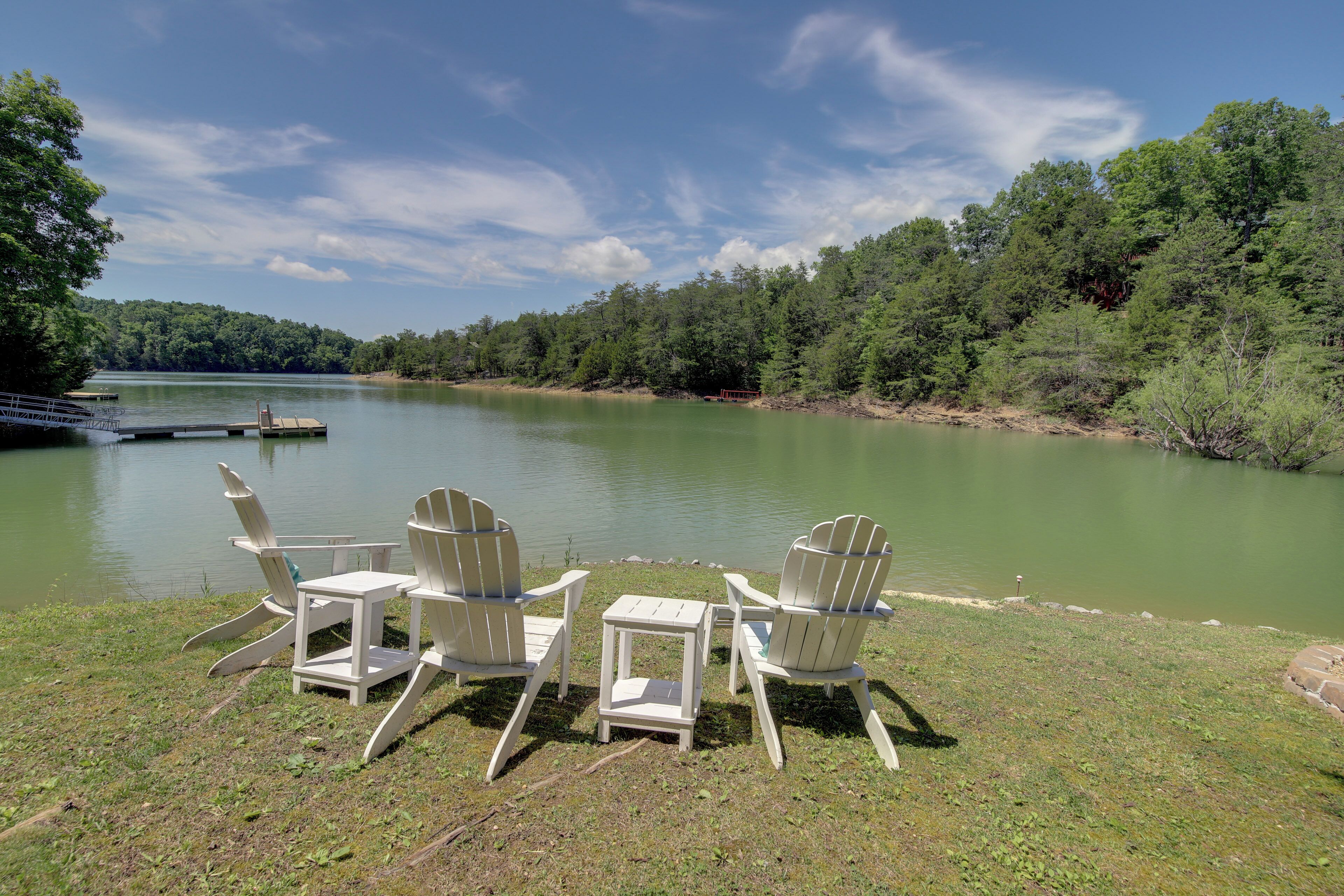 A lakeside view with three Adirondack chairs and a small table on a grassy area.