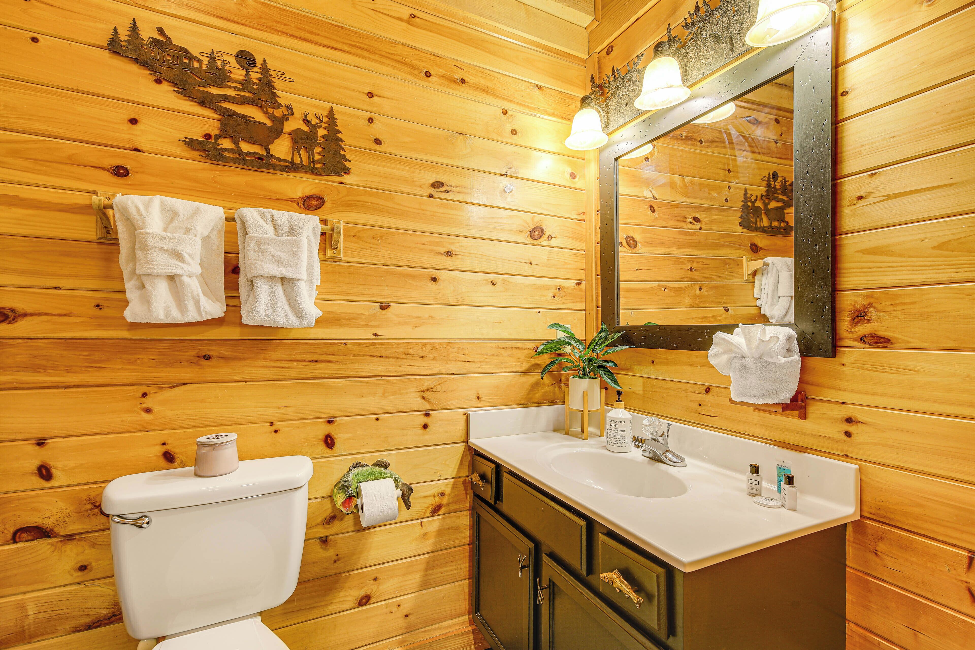 Une salle de bain rustique, aménagée dans une cabane en bois, avec des toilettes, un lavabo et un miroir. Des serviettes sont accrochées au mur et une plante est posée sur le lavabo.
