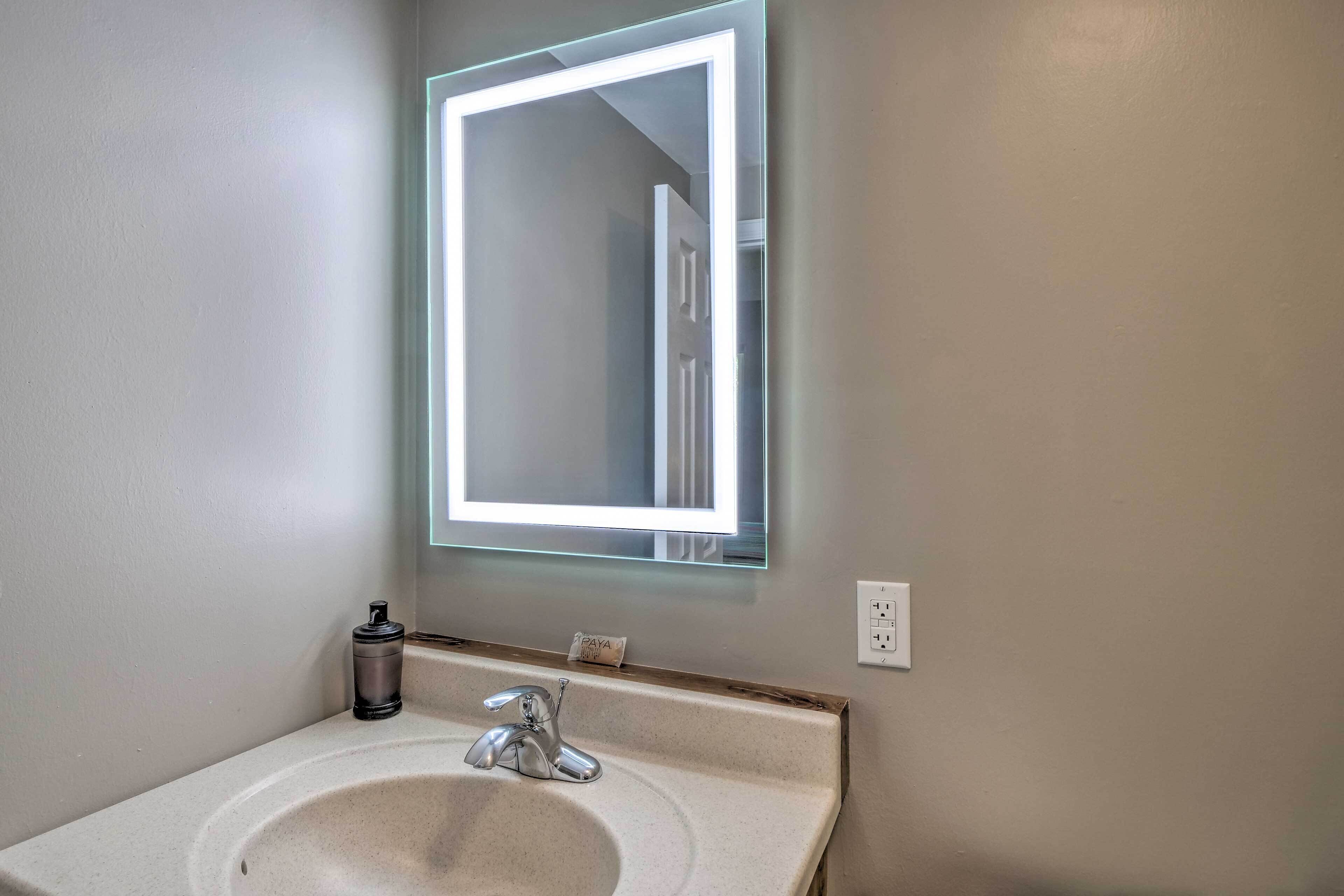 A bathroom with a white sink, a mirror with a light, and a small bottle on the counter.