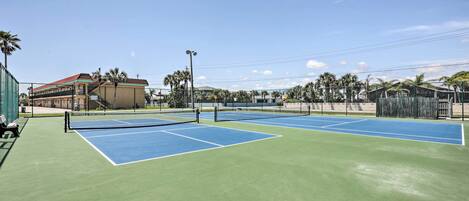 A tennis court with multiple courts, surrounded by a fence, and a building with a red roof in the background.