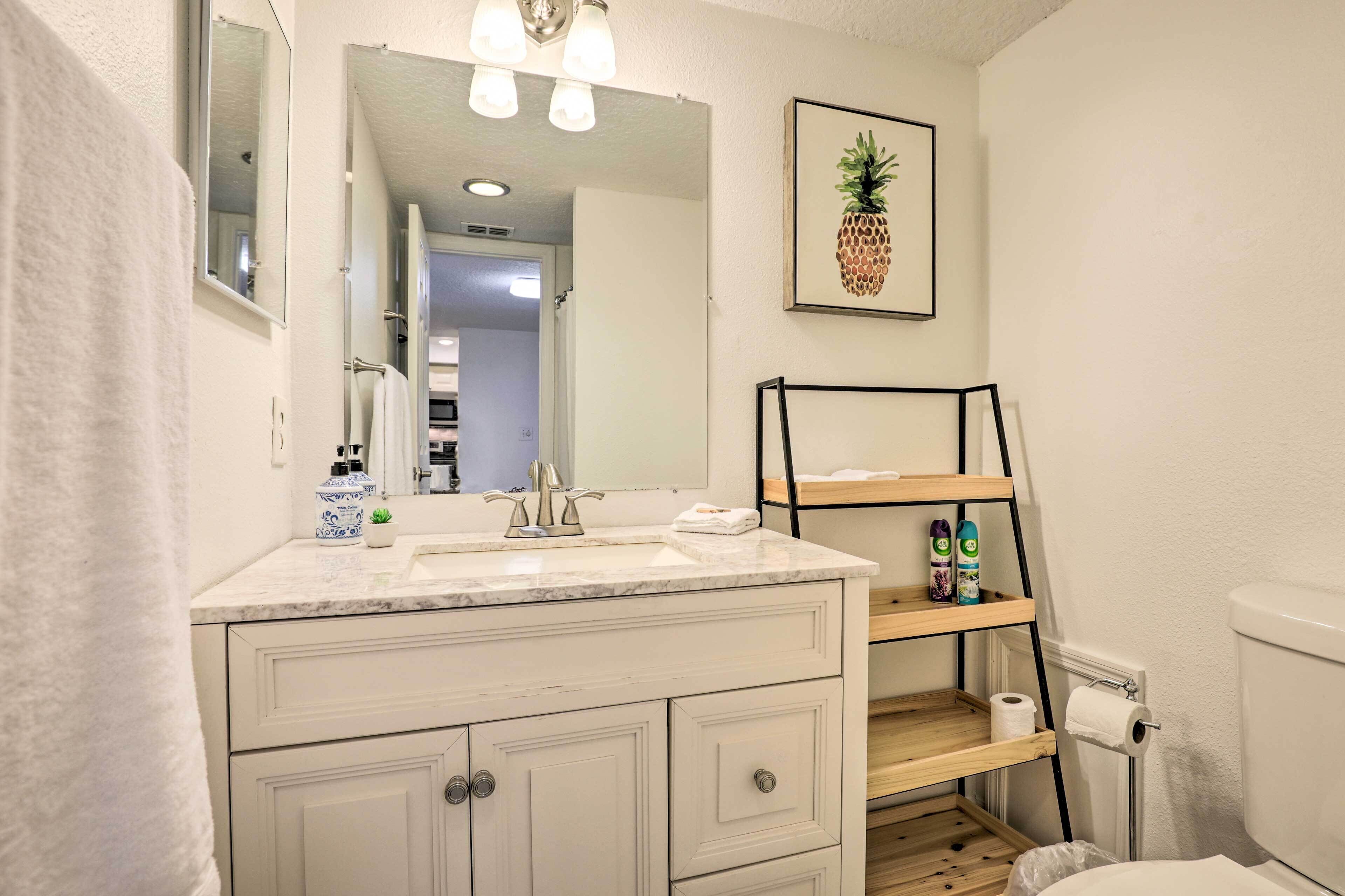 A bathroom with a white vanity, a mirror, a toilet, and a shelf with toiletries.