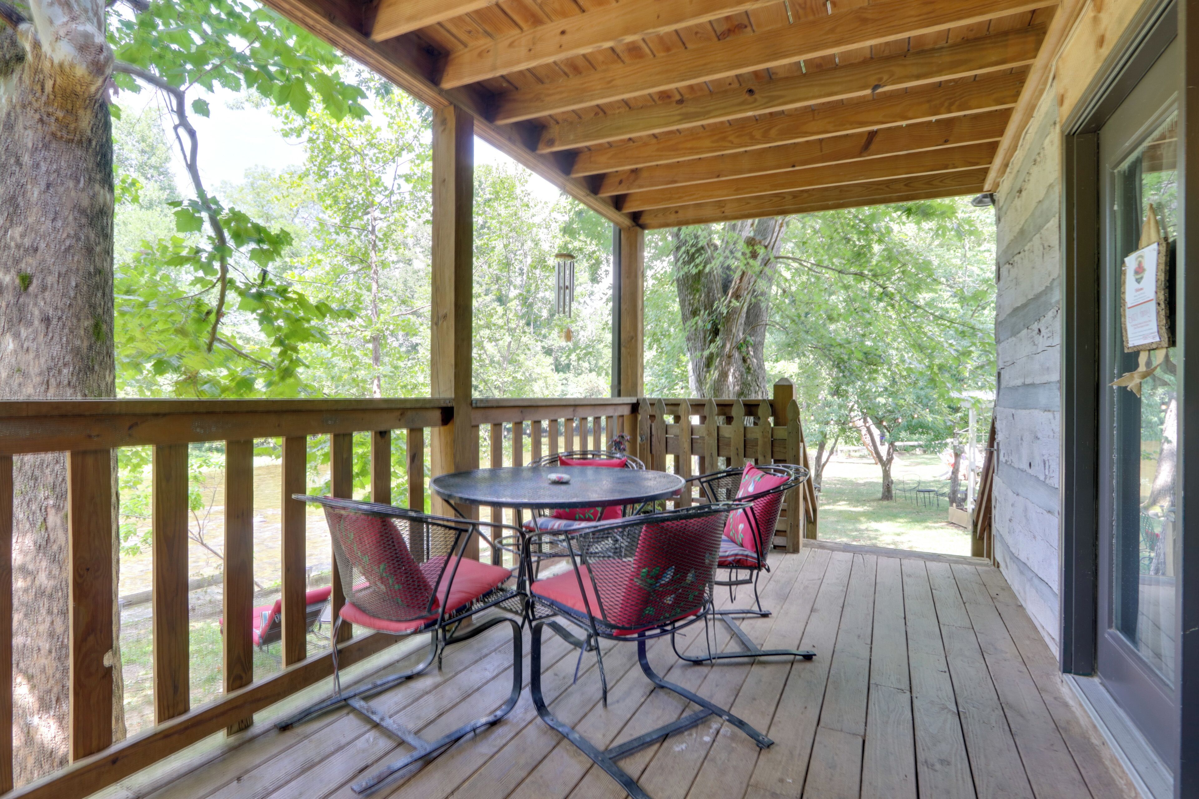 A covered outdoor patio with a wooden table and chairs, surrounded by a wooden railing, overlooking a wooded area.