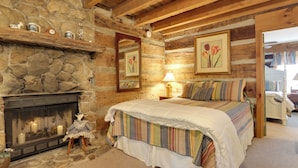Ein gemütliches Schlafzimmer in einer Blockhütte mit Kamin aus Stein, einem Bett mit gestreifter Bettwäsche und einem Nachttisch mit Lampe.
