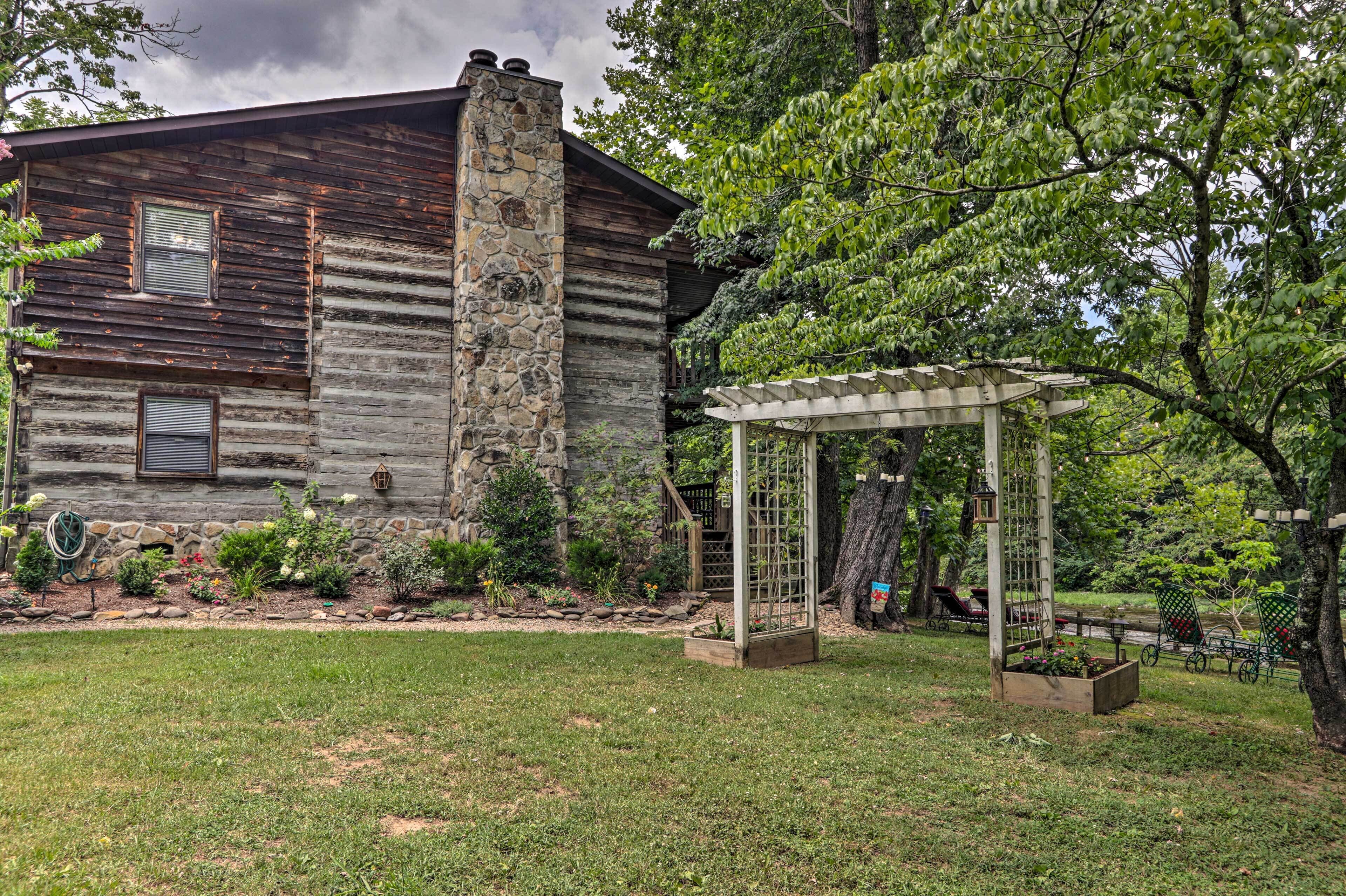A rustic log cabin with a stone chimney, surrounded by a well-maintained garden and a pergola.
