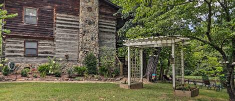A rustic log cabin with a stone chimney, surrounded by a well-maintained garden and a pergola.