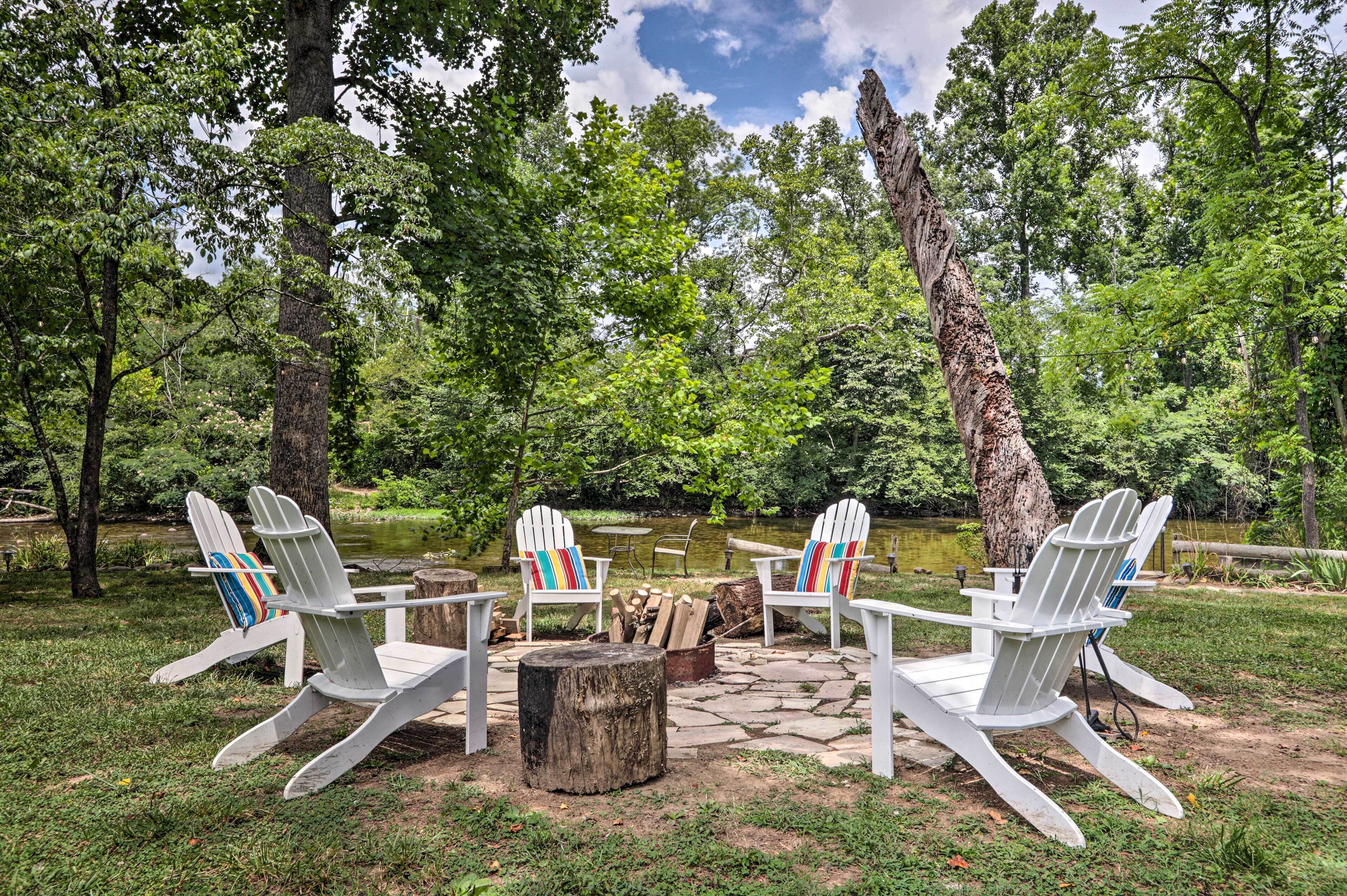 A serene outdoor seating area with Adirondack chairs and a fire pit, surrounded by lush greenery and a pond.