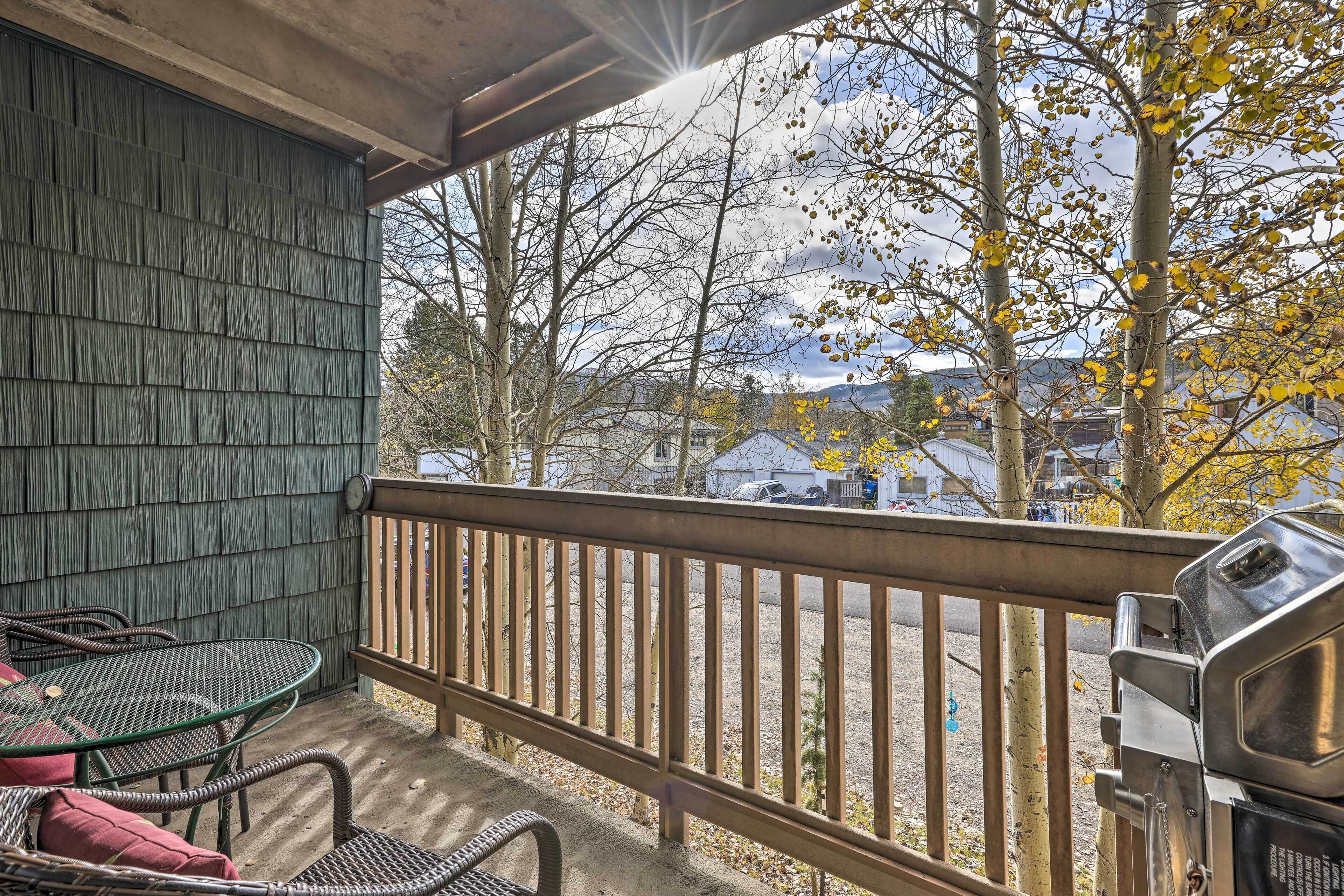 A balcony with a metal table and chairs, a barbecue grill, and a view of trees and houses.