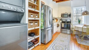 A modern kitchen with a stainless steel refrigerator, oven, and microwave. There are wooden shelves with baskets and a woven chair. - Deck & Fire Pit: Cottage Near Monadnock State Park (Jaffrey)
