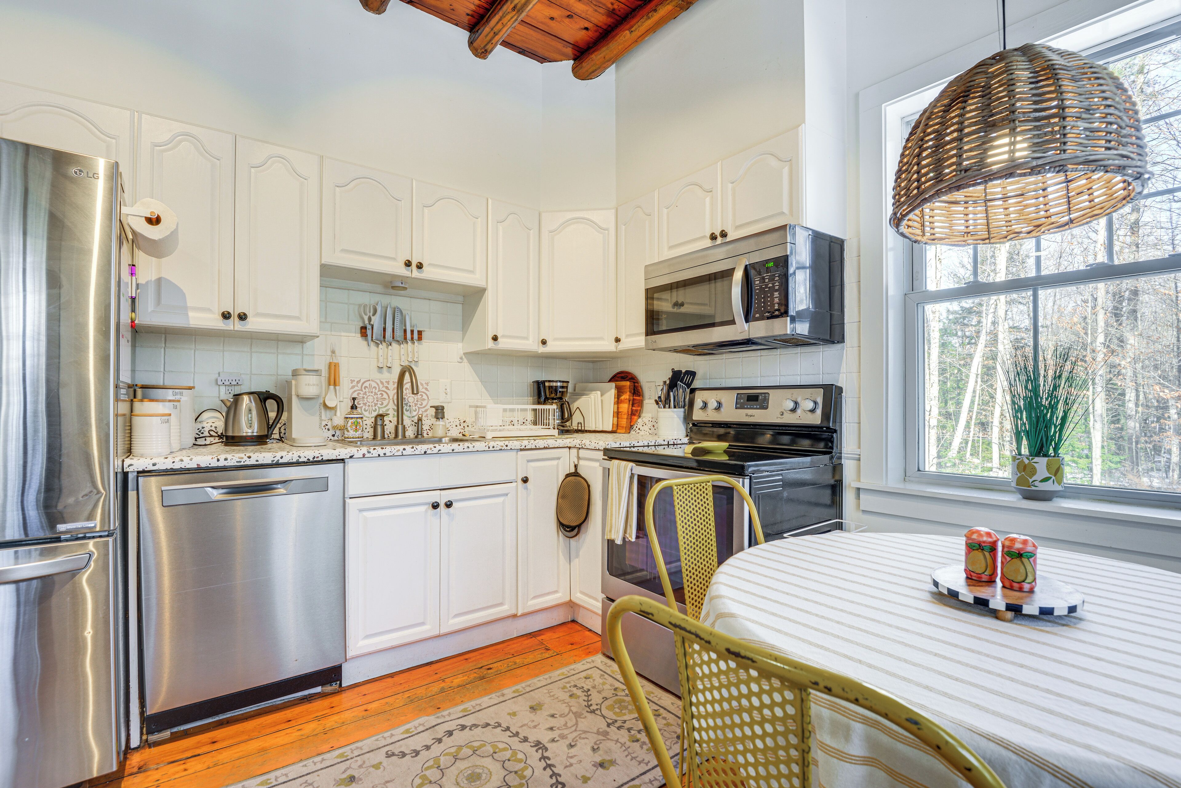 A kitchen with white cabinets, stainless steel appliances, a dining table with a striped tablecloth, and a wicker pendant light.