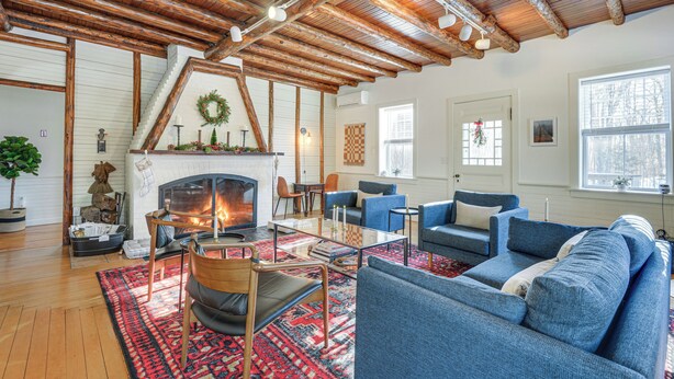 A cozy living room with a fireplace, blue sofas, wooden beams, and a large window. - Deck & Fire Pit: Cottage Near Monadnock State Park (Jaffrey)