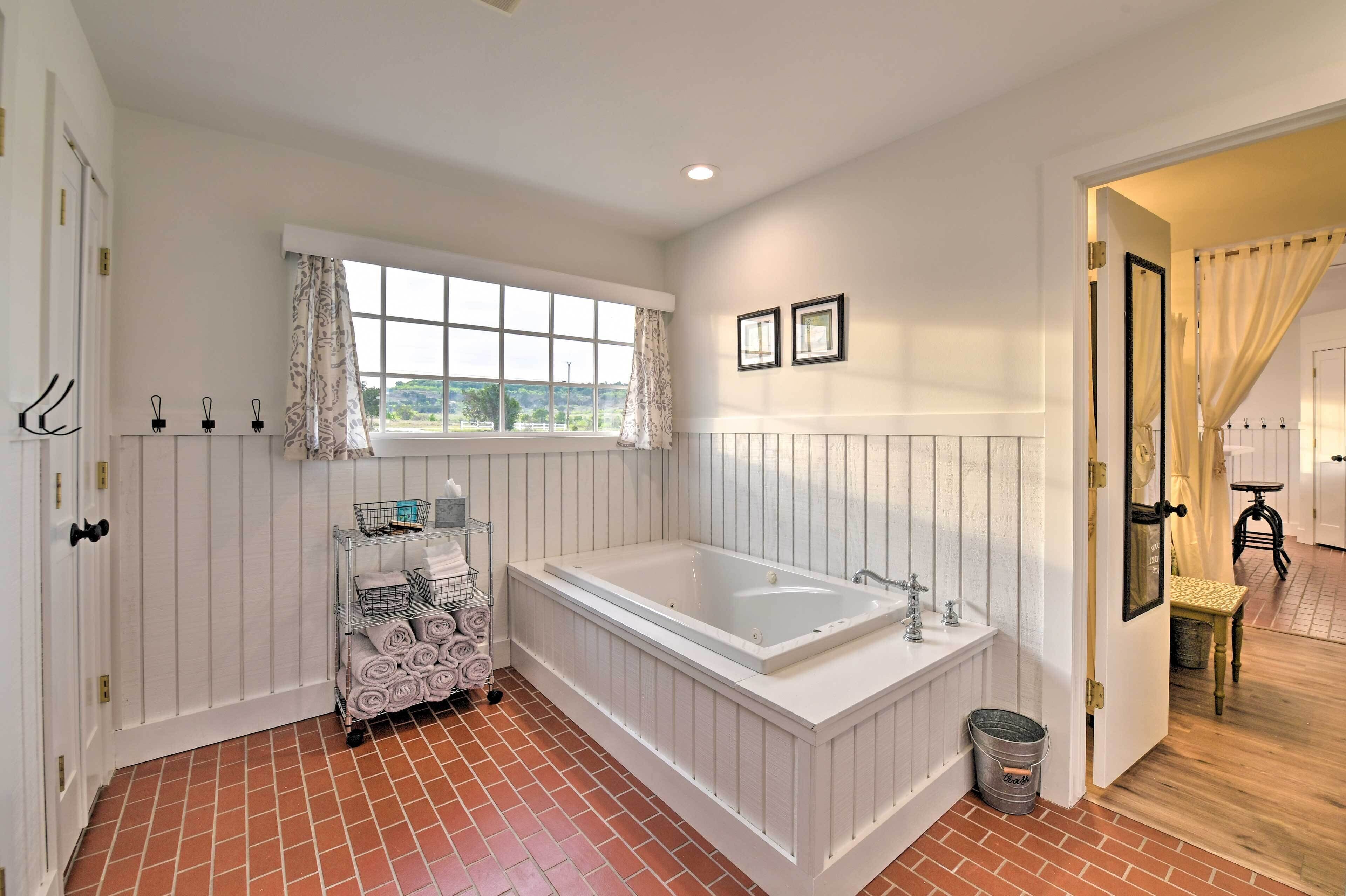 Un baño con una bañera grande, una ventana con cortinas, un perchero y una mesita con una silla.